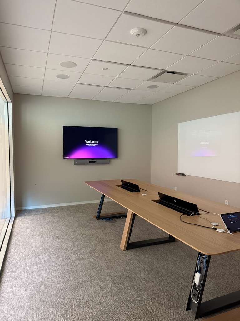 Corner meeting room with a display and video bar, glass whiteboard and pop-up table boxes for power and data