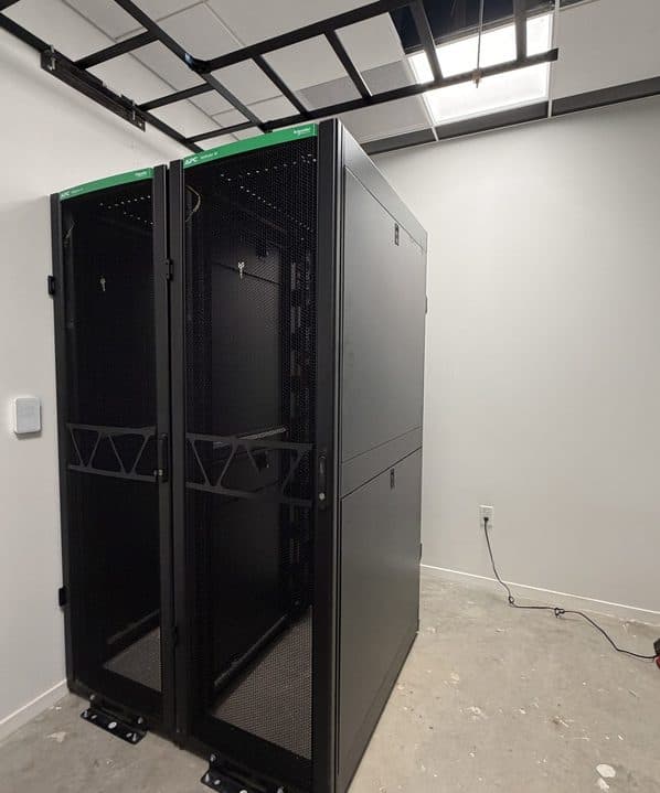 Two black APC NetShelter server racks side by side under an overhead ladder rack