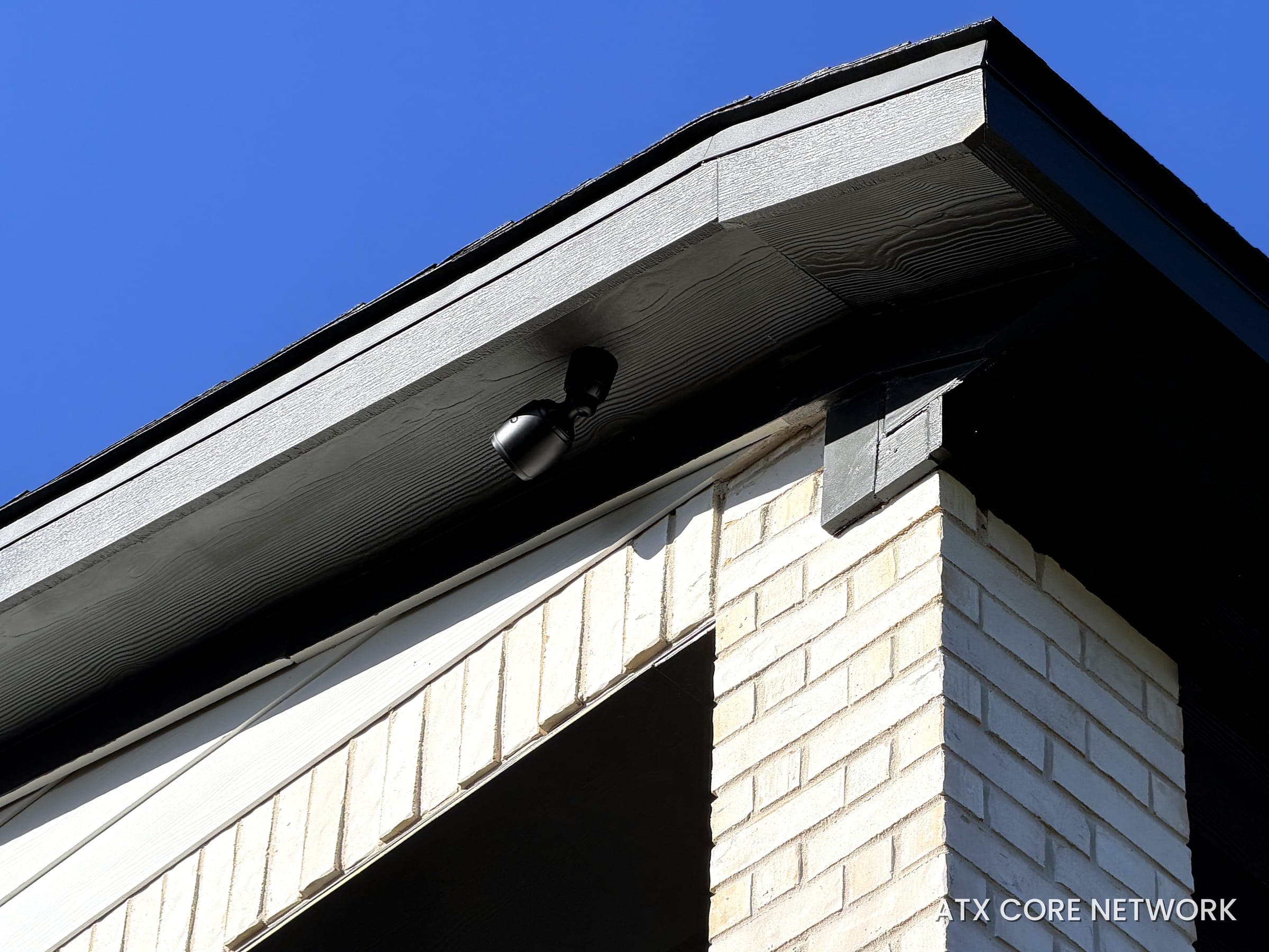 Black bullet IP camera mounted flush under the dark soffit of a modern white-brick home