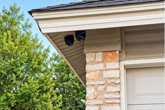 Two black dome IP cameras under the soffit at the corner of a stone home, beside the garage door